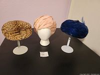 Three vintage hats displayed on mannequins: cheetah print hat, pink feather Christian Dior hat, blue feather hat.