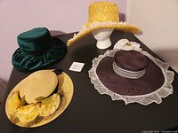 Four wide brim hats arranged on a black surface including yellow with white flowers, green with bow, cream with yellow flower and black ribbon, burgundy/plum with white flowers.