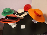 Lot of five colorful women's hats on display stands, showing red, orange, green, cream colors and various brim sizes.