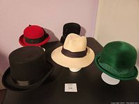 Five vintage hats displayed on table including red bowler, black top hat, beige Panama style hat, green felt bowler-style, and black cloche style