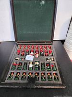 Open wooden box showing organized foam slots filled with Christmas-themed chess pieces in red and green colors.