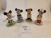 Front view of four Disney ceramic figurines (2 Mickey and 2 Minnie) standing on green bases, showing colorful outfits and character details. Includes size reference with a ruler and label.