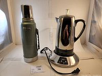 Farberware coffee pot on base with black power cord adjacent to Stanley green thermos with silver trim, both set against white backdrop.