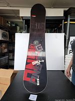 Front and top surface view of GNU Carbon High Beam snowboard showing the graphic and binding insert holes.