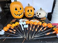 Photo showing three lighted pumpkins of sizes 17", 12", and 8" with jack-o-lantern faces and various lawn stake solar lights with Halloween themes arranged on a surface.