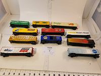 Photo showing 11 different HO scale railroad cars arranged on a white surface with measuring ruler for scale. Various colors and brands identified including Popsicle, NAPA, Coast to Coast.