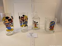 Four glass items in a row on white background: two tall Mickey Mouse glasses, one shorter glass with Mickey head emboss, and a crowned candy jar with Mickey print.