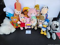 Front view of the entire plush lot showing various Disney, Strawberry Shortcake, Smurf, Sesame Street, Sega characters arranged in two rows on a black surface.