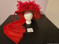 Front angle view of large wide brim red derby hat with attached long red train, mounted on white mannequin head.