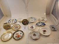 Photo of 12 small decorative plates, bowls, cups, and a vase arranged in a white box with some including floral and gold patterns