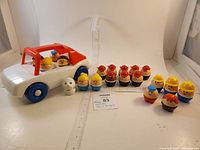 Full view of white and red Little Tikes plastic car with blue wheels, showing 4 figures inside and 16 figures arranged next to car including a white dog figure