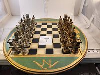Full view of the round chess board with all pieces arranged at starting positions, showing two tones: silver and gold painted figures in Napoleonic style and the painted circular board with gold accents and 'N' symbol.