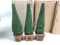 Three ceramic topiary decor pieces displayed with green artificial foliage and terracotta-colored bases with fleur-de-lis motif, set before cardboard boxes.