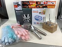 Overview of kitchenware lot showing reusable grilling sheets, electric lunch box, silicone gloves, garlic press, food grater, rotisserie forks, and UNiWEIGH digital scale.