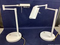 Front view of both white metal bedside lamps showing the circular bases, vertical supports, and adjustable swing arms.