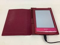 Sony e-reader device with pink bezel and physical buttons displayed open in red protective cover, connected to charging cable.