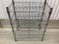 Front view of the stainless steel wire wine rack showing three adjustable shelves and four vertical poles, placed against a white curtain background.