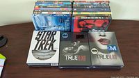 Photo showing a collection of DVD movies arranged in two stacks plus two unopened True Blood series box sets and a Star Trek set.