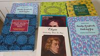 Seven music books arranged side by side, showing diverse covers with floral patterns, blue and green backgrounds, and portraits of Chopin and Beethoven.