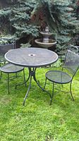 Full view of the round metal mesh bistro table on grass with two matching metal mesh chairs, black powder-coated metal.