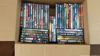 Top-down view of a cardboard box filled with multiple DVD cases showing various movie titles doubled stacked