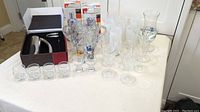 Wide view of assorted clear glassware including wine glasses, goblets, and shot glasses arranged on a white surface next to boxed cocktail shaker and ice bucket.