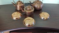 Three vintage copper kettles and three decorative copper wall hangings displayed on a dark wooden table.