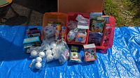 Photo showing plastic bins filled with assorted craft supplies such as white foam balls, stickers, foam shapes, beads, and glue tubes arranged on a blue tarp.