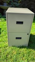 Front view of beige two drawer metal filing cabinet standing upright outdoors on grass.