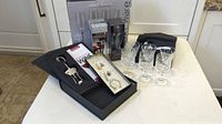 Photo showing full lot on white table, includes boxed wine decanter aerator, deluxe corkscrew opener in black foam box, small booklet, set of wine charms, 4 wine flutes, 6 etched wine glasses