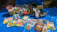 Wide shot of all items including piles of activity books, paint sets, glue sticks, and small containers with various craft supplies and party favors, arranged on a blue tarp outdoors.