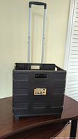 Front view of the closed foldable black plastic crate with extended silver telescoping handle and black grip standing on a table.