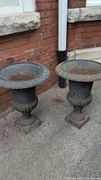 Pair of tall metal urns outdoors on concrete next to brick wall showing full view