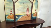 Full view of three wooden panels painted with a seaside scene including lighthouse, rocks, sailboats, and sky. Panels have arch tops and metal hinges.