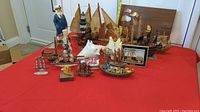 Wide view of multiple ship models in various materials including wood, metal, amber, and shells on a red surface.
