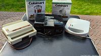 Full view of five small appliances: Sunbeam sandwich maker, black toaster oven, white sandwich grill, and two unopened Cuisinart boxes (juice extractor and food processor).