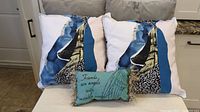 Five throw cushions arranged on a couch: four large square cushions with abstract blue, black, white, beige design, and one smaller mossy green rectangular cushion with text and fringed edge.