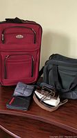 Two carry-on bags (one red wheeled suitcase, one black shoulder bag), a passport holder, a money belt, and a travel iron placed on a wooden surface.