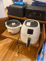 Philips air fryer and Krups crockpot shown side by side on wooden stairs, also includes a black printer and another appliance in background.