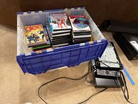 Photo showing a plastic storage bin filled with DVDs including 'Glee' and other family-oriented titles, alongside two black Sony Blu-ray players and a remote control on the floor.