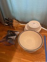 Knives housed in a wooden block, ringed ceramic serving bowls, and a stack of ceramic plates on hardwood floor.