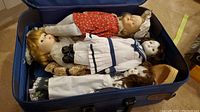 Five vintage porcelain dolls lying in a blue suitcase, showing varied clothing and hairstyles.