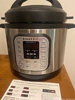 Front view of stainless steel Instant Pot pressure cooker with control panel visible showing various cooking functions and digital display. Lid is closed with black handle.