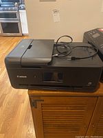 Front and top view of Canon TS9520 printer with power cord placed on top, showing LCD screen and paper trays.