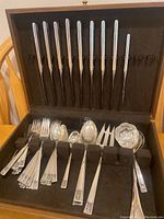 Wood and felt-lined wooden box open showing 11 knives in top rack and assorted forks, spoons and carving fork on bottom wedge.
