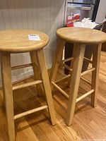 Two wooden bar stools shown side by side, displaying round seats and four legs with support bars. Natural wood finish is visible with some staining and wear.