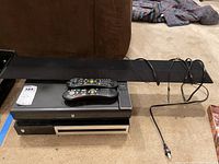 Photo showing the two TiVo DVR units stacked, each with a remote on top, and the Vizio sound bar placed behind. Items rest on carpet flooring.