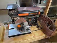 Front and side view of Wen 4x36 belt sander and 6 inch disc sander on wooden surface with sanding belts stacked next to it.