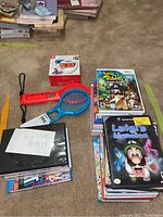 Accessories and game stacks on carpet showing two tennis racket shells and one boxed Joy-Con wheel.