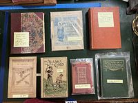Seven vintage books laid out showing titles and condition. Includes 'Gold Hunting in Alaska,' 'In Alaska,' 'To Alaska for Gold,' poetry collections and others with worn covers and vintage design.
